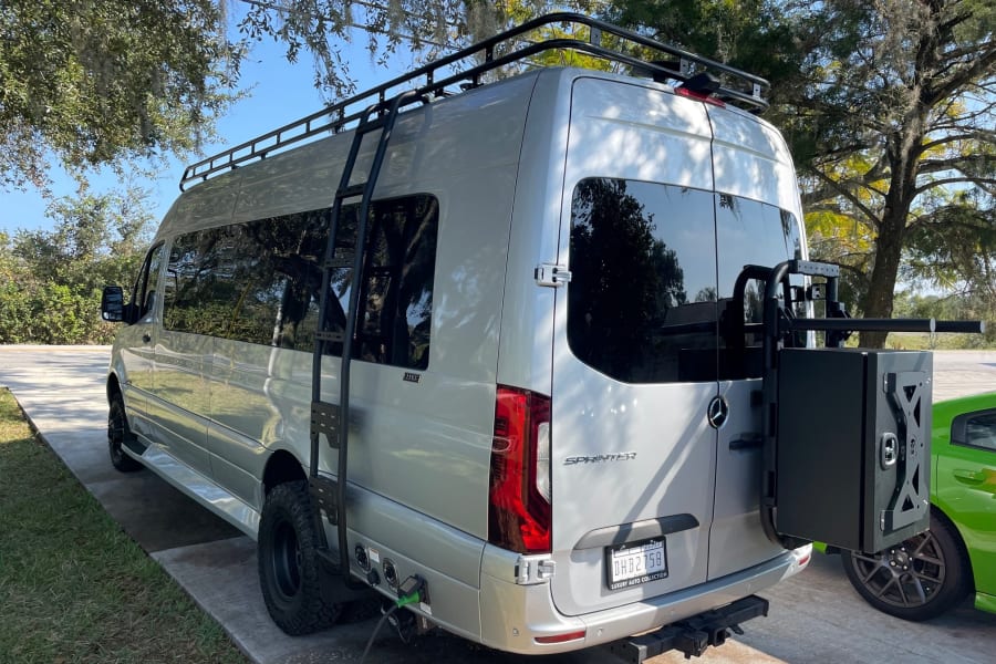 This silver Mercedes Sprinter rv rental showcases a roof rack, ladder, and rear storage box, ideal for travel.