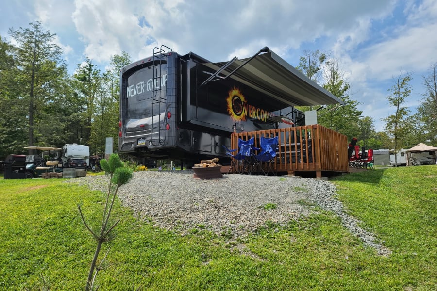 An RV rental is set up on a spacious gravel campsite, featuring a wooden deck, extended awning, and fire pit.
