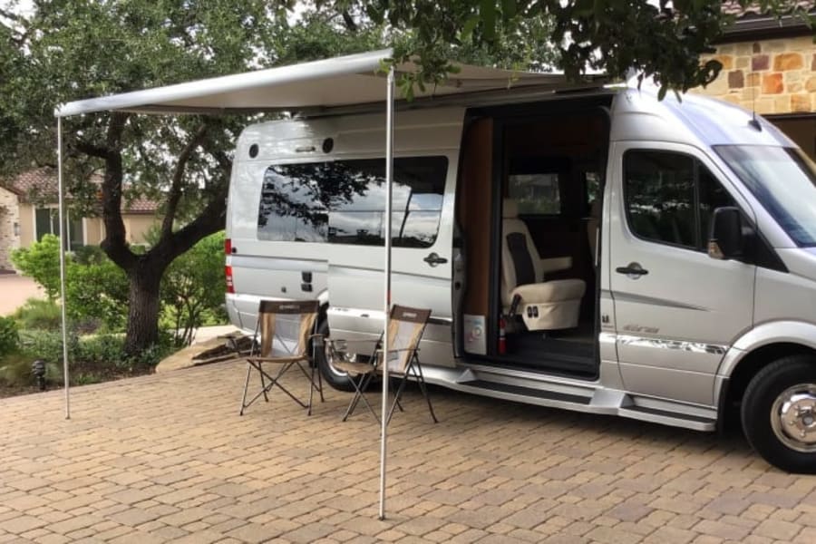 A silver Mercedes-Benz Sprinter RV rental with its awning extended, parked on a paver patio with chairs. Ideal for travel.