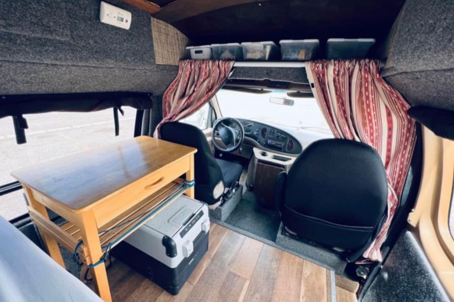Cozy RV rental interior with wooden table, portable fridge, and driver's cabin. Ideal camper van setup.