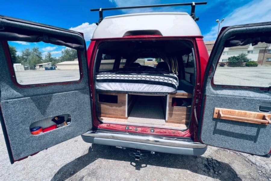 Red high-top camper van for rv rental, showing custom interior with bed and storage from the open rear doors.