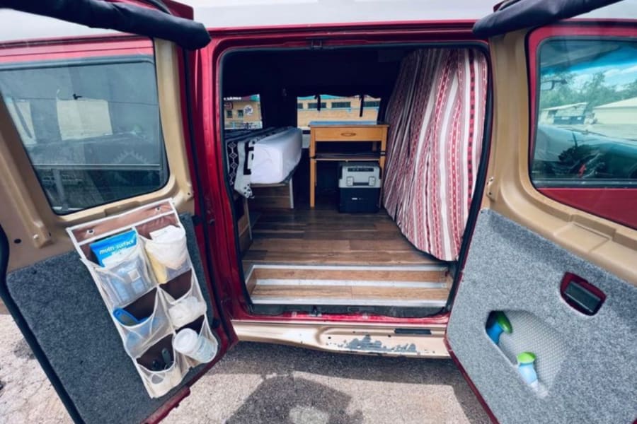 Step into this RV rental! Converted van interior features a bed, desk, cooler, & smart storage solutions.