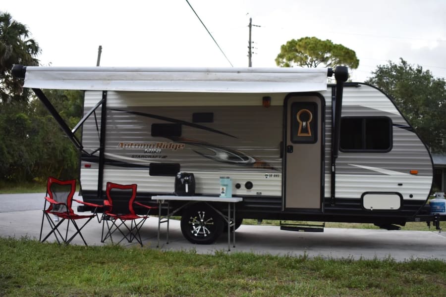 This Autumn Ridge Mini RV rental features an extended awning, two red chairs, and a table with appliances for outdoor enjoyment.