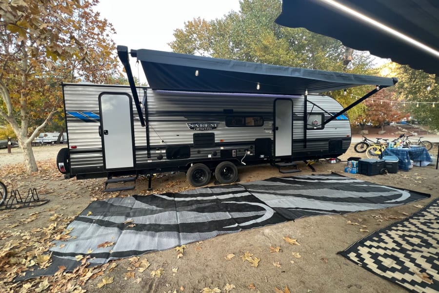 Enjoy fall camping! This RV rental travel trailer is set up at a leafy campsite with an extended awning, outdoor rugs, and bikes.