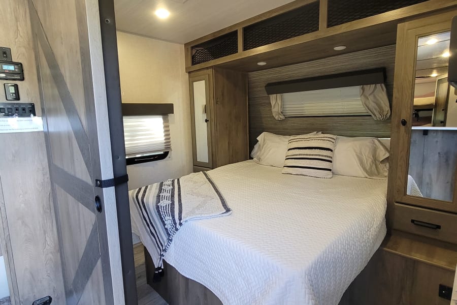 Bright RV rental bedroom with a white quilted bed, wood-toned cabinetry, overhead storage, and a stylish sliding barn door.
