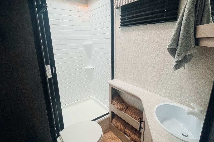 Modern rv rental bathroom featuring a compact shower, toilet, sink vanity with towel storage, and skylight. Clean and functional.