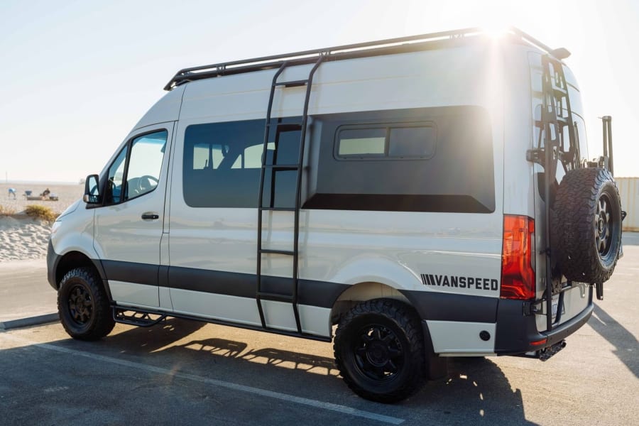 A rugged white VANSPEED adventure van with off-road tires and gear, parked by a sunny beach, ready for an RV rental journey.