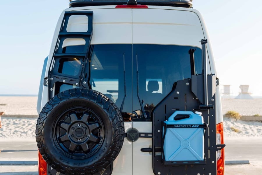 Rear view of a custom white RV rental van, equipped with a ladder, spare tire, and utility can, parked by the beach.