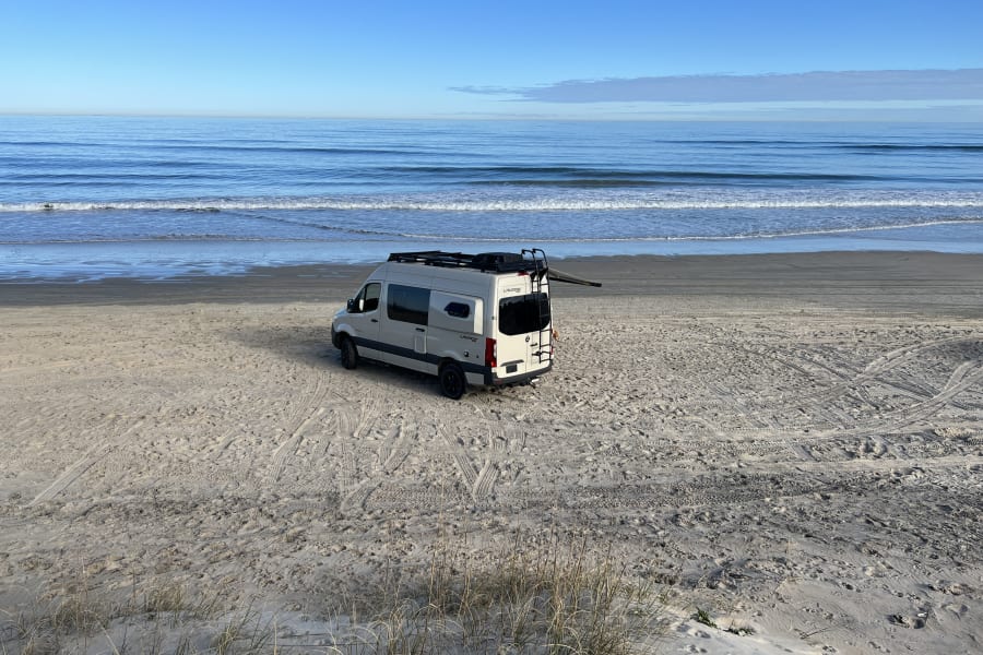 White RV rental camper van parked on a sandy beach, ocean waves rolling in under a clear blue sky. Ideal beach trip.