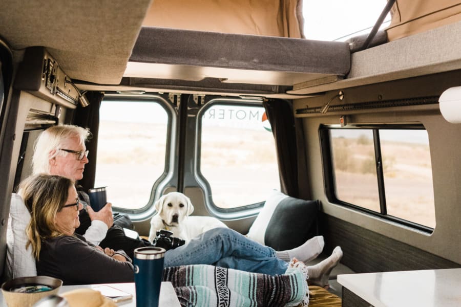 Inside a spacious Moterra rv rental campervan, two people and a dog relax, enjoying drinks and desert views.