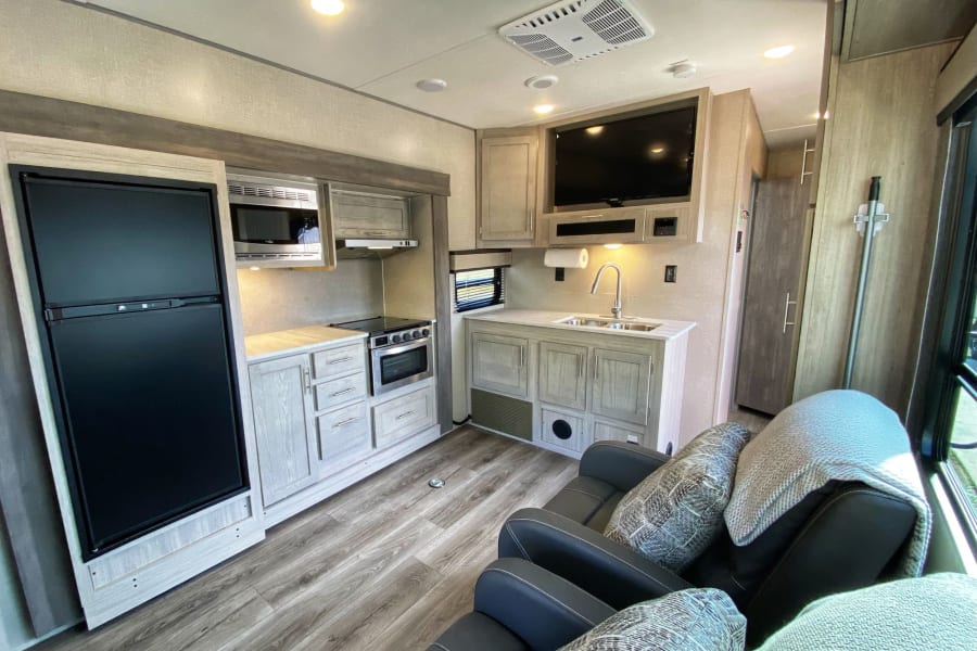 Interior of an RV rental showing a modern kitchen with light cabinets, fridge, stove, sink, and comfy recliners.