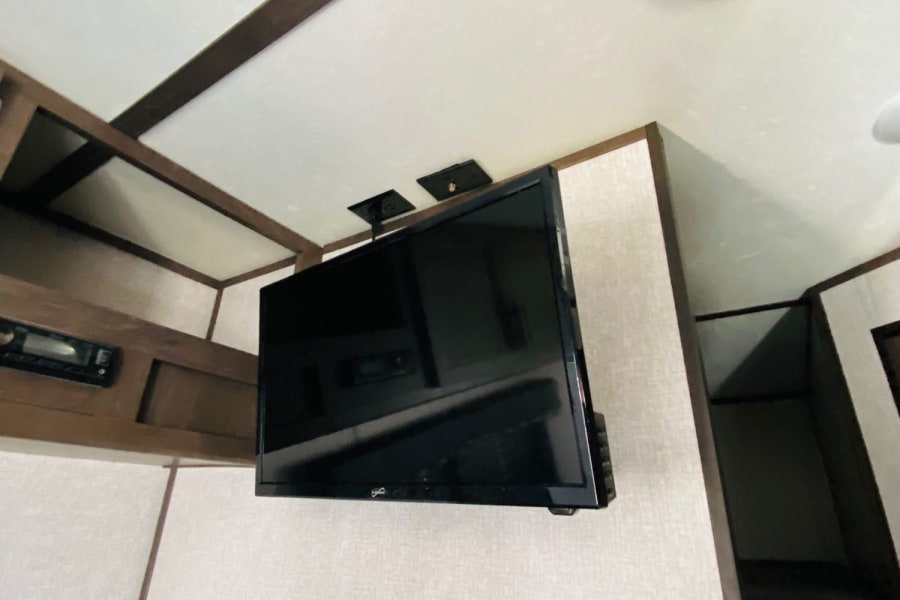 RV rental interior: TV mounted near ceiling, overhead storage, speaker, and control panel. Comfortable bunk area visible.