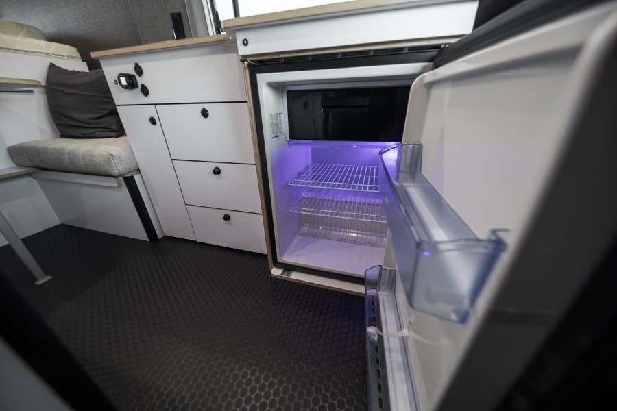 Modern RV rental interior: open fridge with purple light, white cabinets, and comfortable bench seating.