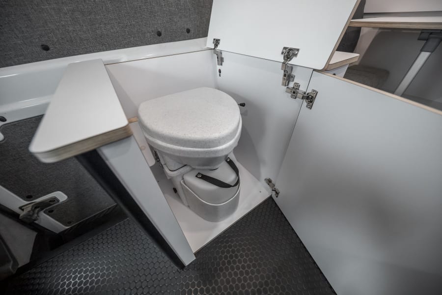 RV rental interior showcasing a compact, white composting toilet with open cabinet doors on a dark, textured floor.
