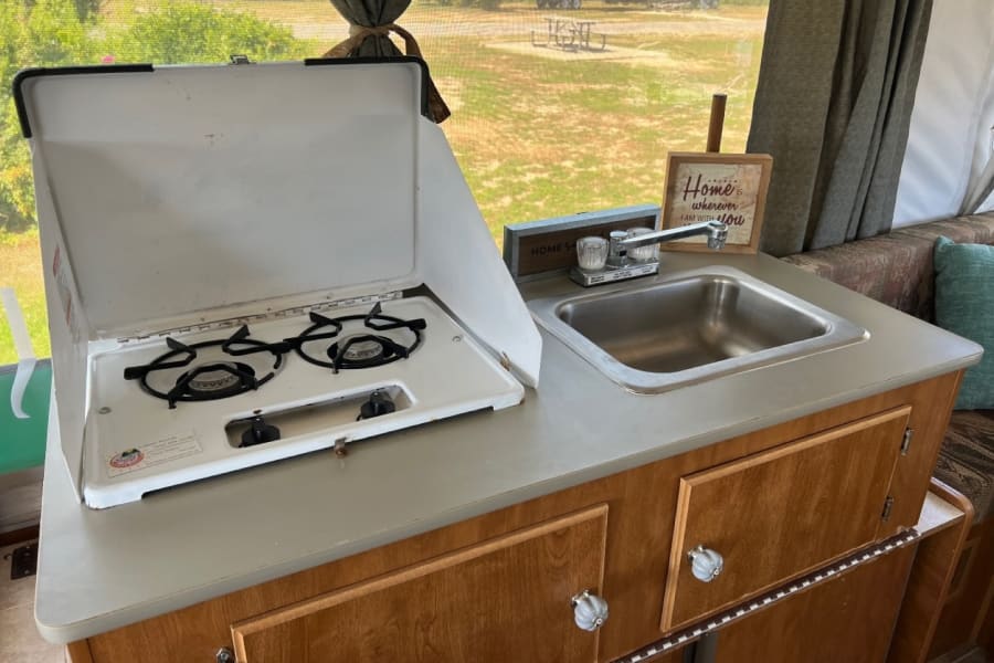 RV rental kitchen: Interior view of light gray counter with two-burner stove and sink, overlooking a scenic campground setting.