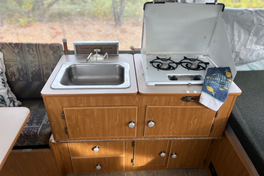 This RV rental features a cozy kitchen with a stainless steel sink, two-burner stove, and wooden cabinetry. Ideal for camping trips.