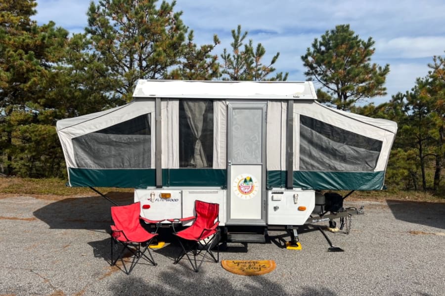 This cozy RV rental pop-up camper is set up at a sunny campsite with red chairs and a fun doormat, perfect for outdoor adventures.