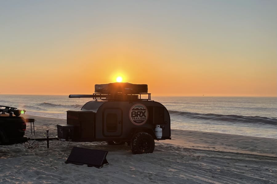 Sunrise beach camping with a BRX teardrop RV rental, a compact trailer parked on sand beside the ocean, a solar panel nearby.