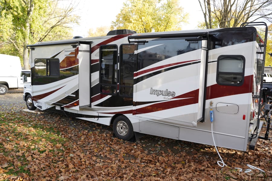 This white, black, and maroon motorhome, a spacious RV rental with extended slide-outs, is parked among autumn leaves.