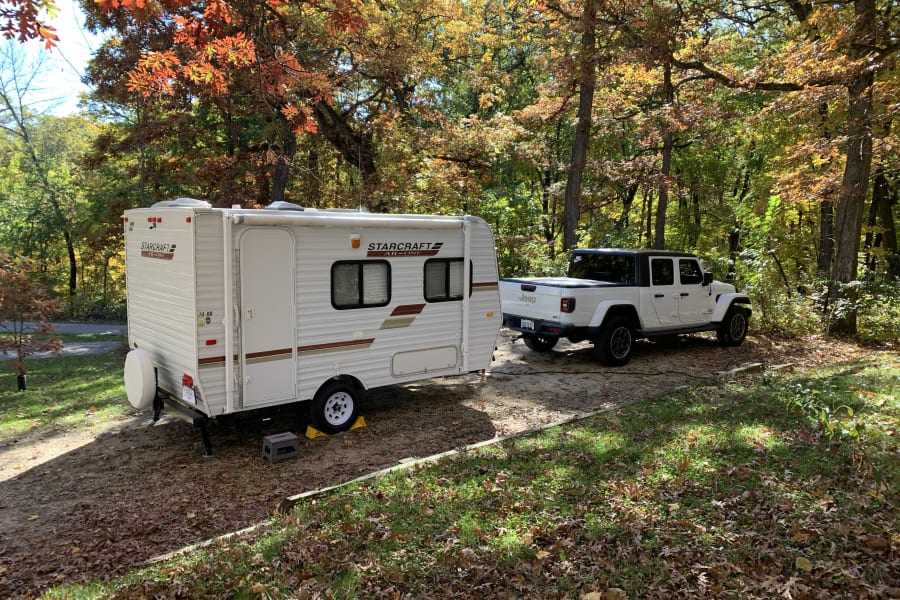 An RV rental and white Jeep Gladiator are parked at a serene autumn campsite, surrounded by trees with vibrant fall foliage.
