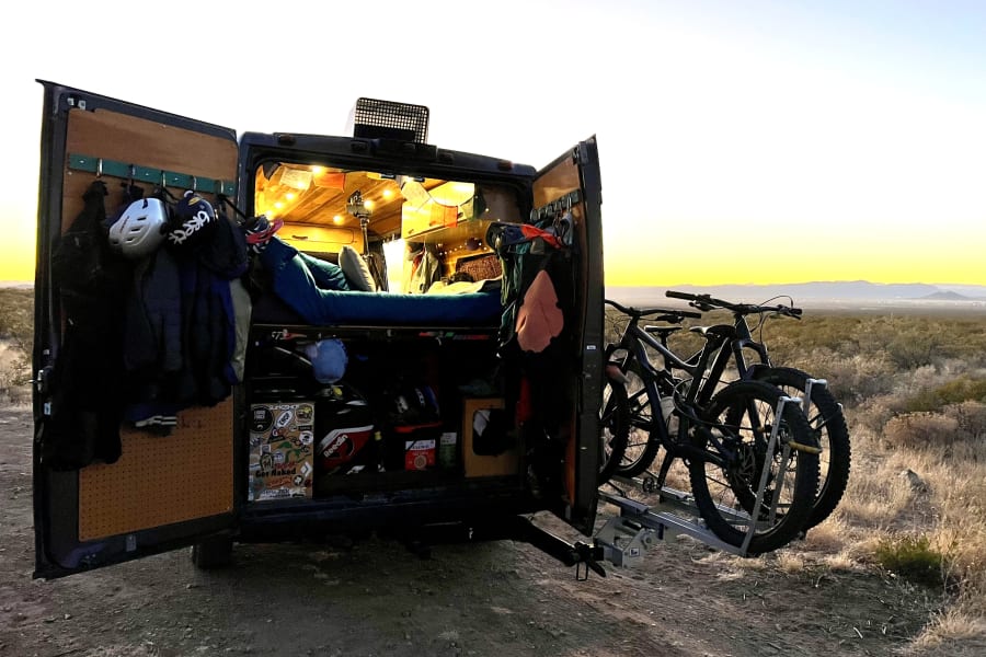 This RV rental camper van, with open rear doors, reveals a cozy interior, bed, gear, and mountain bikes on a hitch rack at sunset.