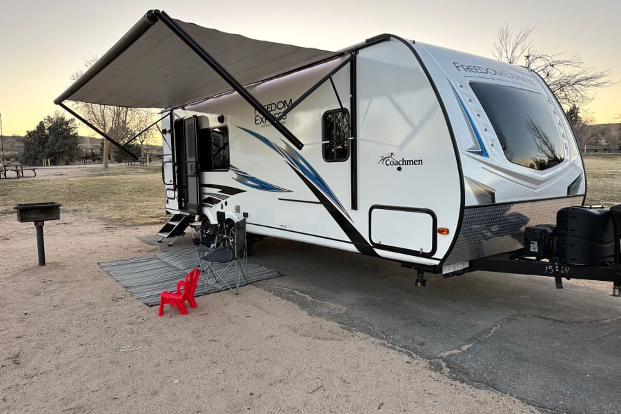 Cozy RV rental set up for camping at sunset, featuring an extended awning, chairs, and a grill.