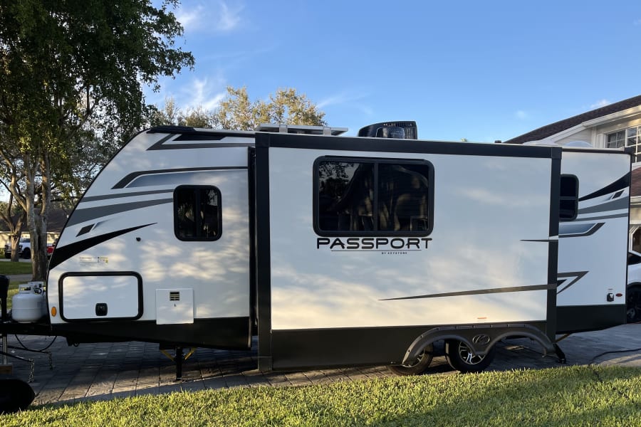 White Passport travel trailer, a spacious RV rental, parked on a driveway with slide-out extended, ready for adventures.