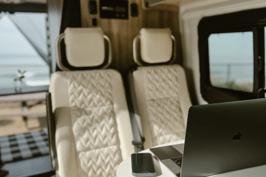 Comfortable RV rental interior with light-colored, quilted seats, a laptop, and phone on a small table. Ocean view outside.