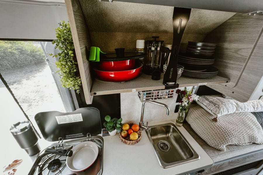 Cozy RV rental kitchen featuring a compact stove, sink, organized shelves with dishes & cookware, and a comfy sleeping area.