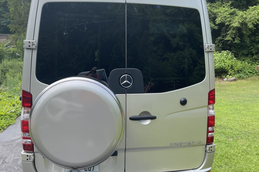 Rear view of a silver Mercedes-Benz Sprinter RV rental, with a spare tire & "WEEKENDER" badge, parked on an asphalt drive.
