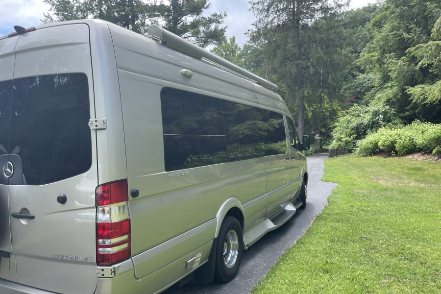 A silver Mercedes-Benz Weekender RV rental is parked on a shaded driveway with lush green grass and tall trees.