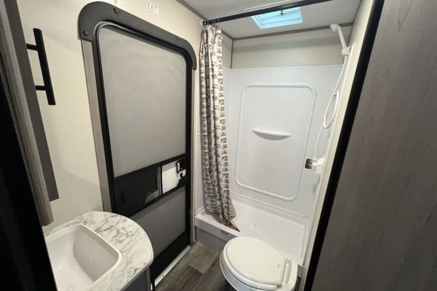 Inside a modern RV rental: a compact bathroom featuring a full shower, toilet, sink, and skylight overhead.