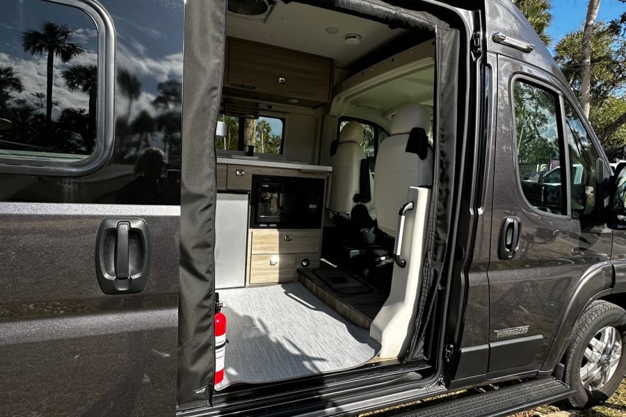Open side door of an RV rental reveals a cozy interior with kitchen, seating, & fire extinguisher. Parked by palm trees.