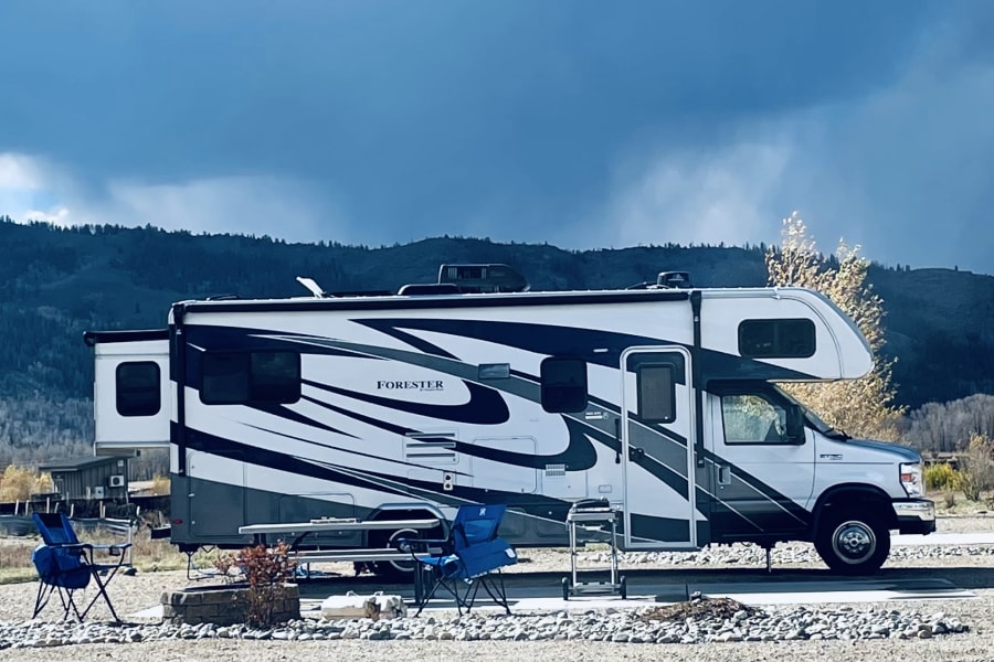 Large white and gray rv rental parked at a gravel campsite with blue chairs, a table, and fire pit. Mountains under a cloudy sky.