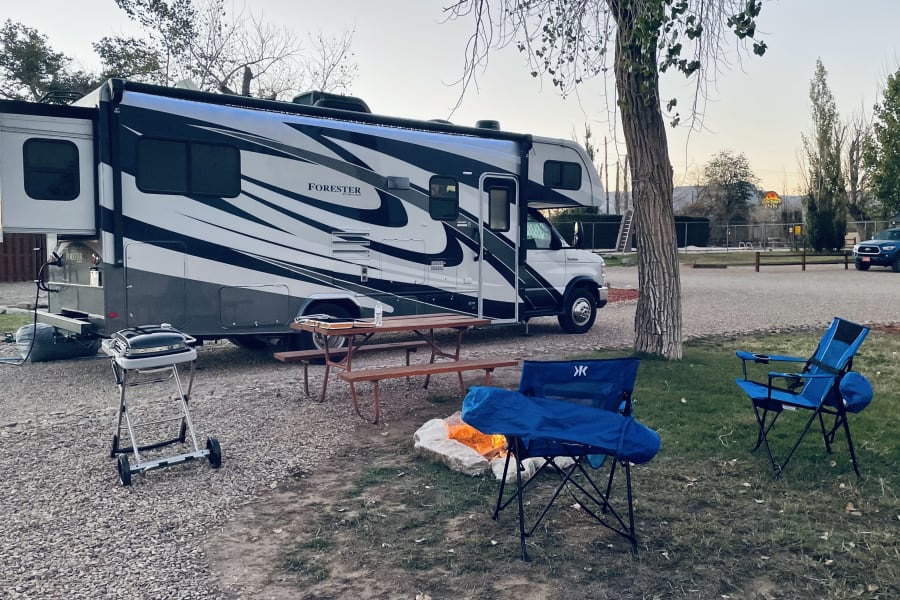 RV rental campsite setup: cozy campfire, grill, and picnic table by a large motorhome for an inviting outdoor experience.