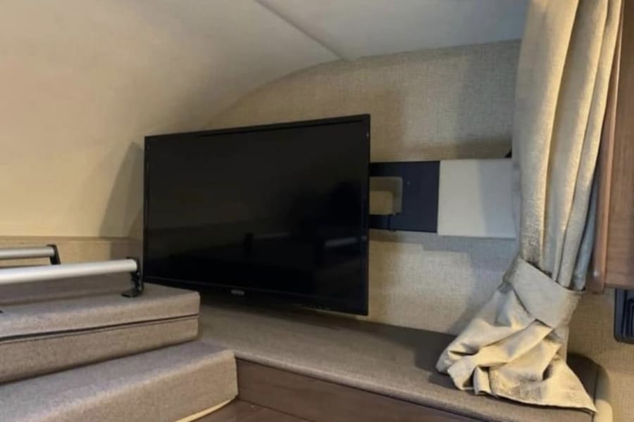 Spacious RV rental interior with a wall-mounted TV over carpeted steps leading to an elevated bunk. A curtain is on right.