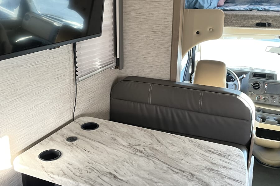 Modern RV rental interior with dinette, marble-look table, TV, and an overhead bunk bed above the cab area.