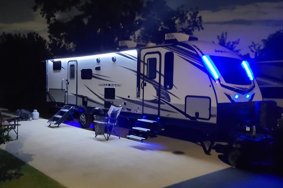This illuminated RV rental travel trailer shines at night with vibrant blue LED lights at a peaceful campsite.