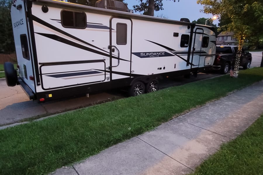 White Sundance Ultra Lite RV rental, a travel trailer, parked on a paved area next to a sidewalk and grass.