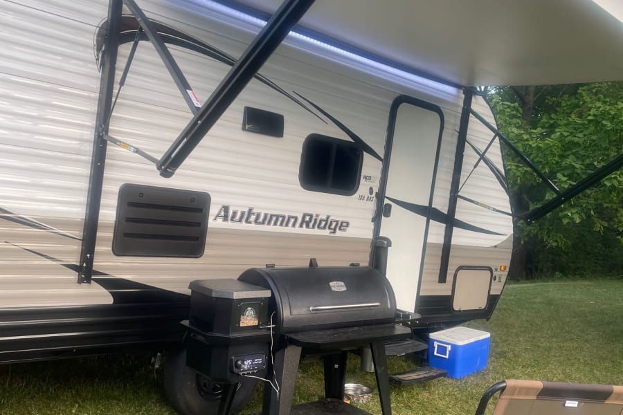 Perfect RV rental camp: travel trailer with illuminated awning, Pit Boss grill, cooler, and relaxing chair.