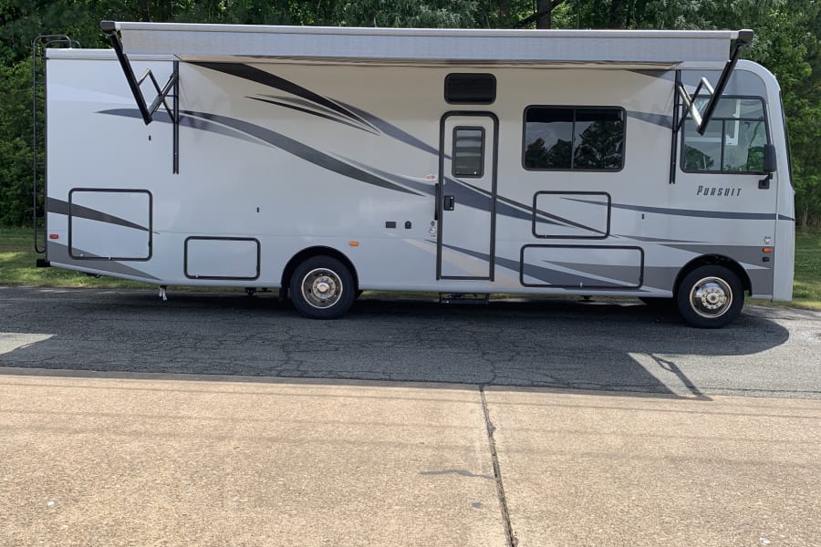 This light-colored Pursuit RV, featuring an extended awning, is parked on pavement against a green tree line, available for rental.