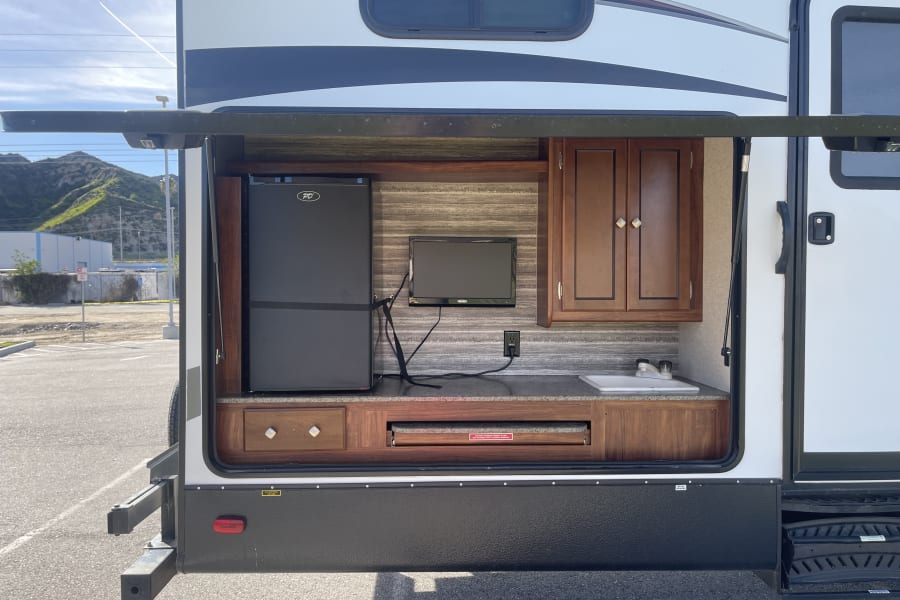 RV rental with an open side panel revealing a fully equipped outdoor kitchen: fridge, TV, sink, & countertop grill.