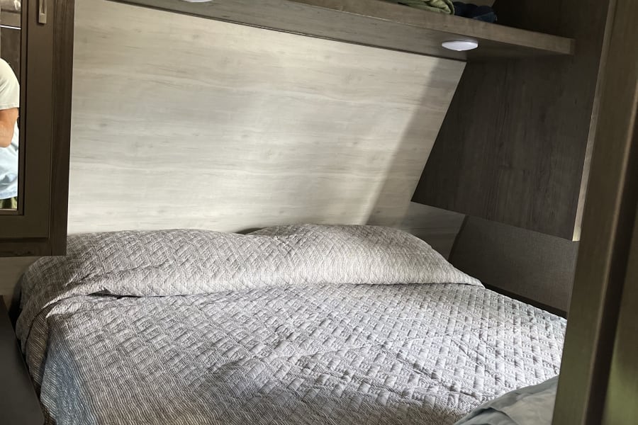 An RV rental bedroom featuring a grey quilted bed, overhead shelf with blankets, and cabinet with mirror.