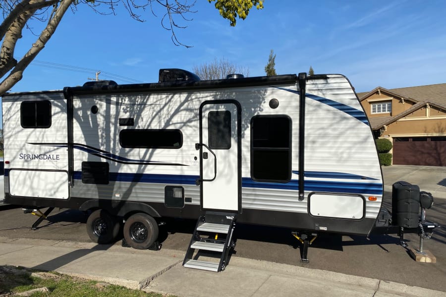 A white and blue Springdale travel trailer, perfect for an RV rental. Parked with steps down under tree shadows.