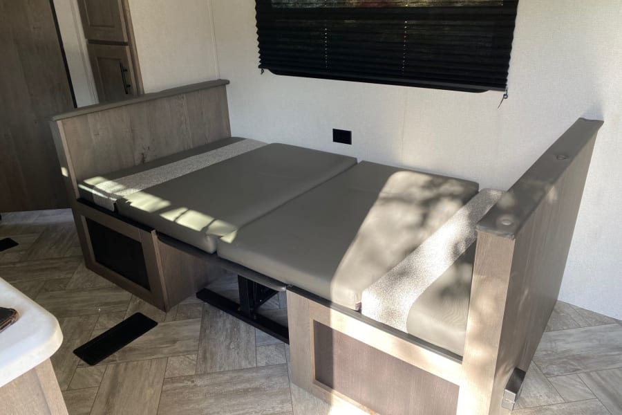 RV rental interior: Wood-toned dinette with gray cushions converted into a bed, bathed in sunlight.