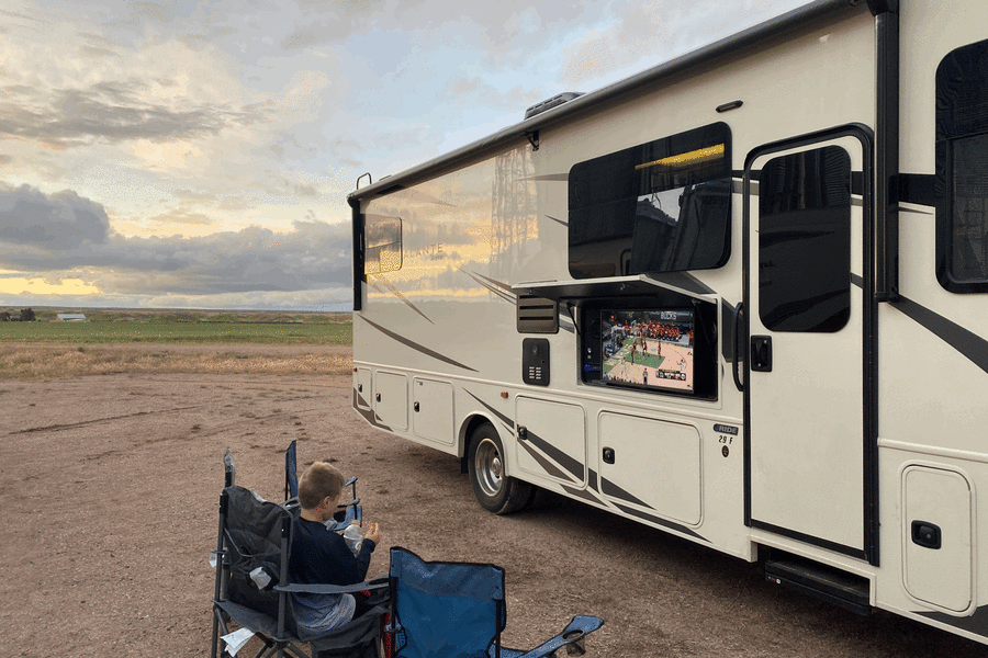 An RV rental provides outdoor entertainment, as a boy watches basketball on its pull-out TV amidst a scenic, open landscape.