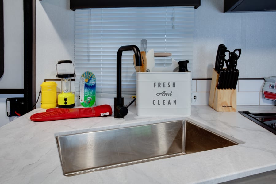 A neat RV rental kitchen workspace: white counters, a stainless steel sink, black faucet, and organized camping-themed essentials.