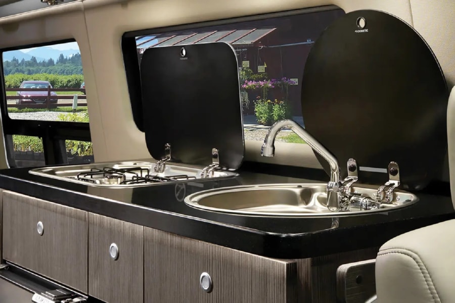 Compact Dometic kitchen inside an rv rental, featuring a stove, sink, and storage units, with a serene outdoor vista.