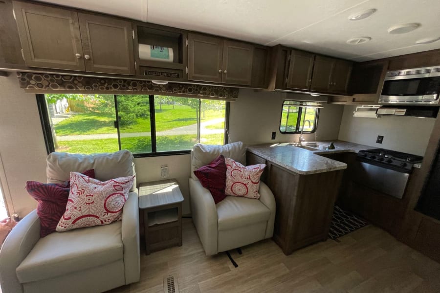 Inside this RV rental: Two comfy armchairs with pillows, a side table, and a compact kitchen area with stove and microwave.