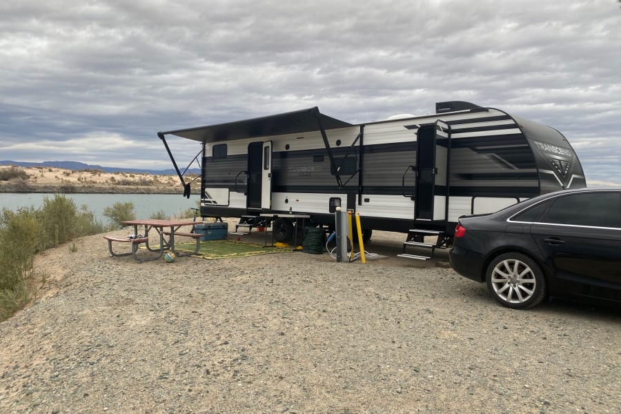 Spacious RV rental parked by a river, featuring an extended awning, picnic table, and utility hookups on a cloudy day.